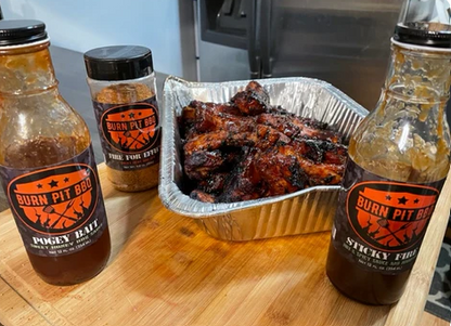 Three bottles of 'Burn Pit BBQ' barbecue sauce on a wooden surface with a container of ribs.